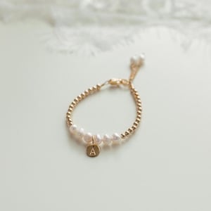 Op de afbeelding: Een gouden armband met een delicate ketting en kleine parels. Een gouden bedel met de letter 'A' hangt aan de armband.