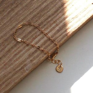 May include: Gold-colored bracelet with a delicate chain design. The bracelet features a small, round charm with the letter "S" on it. The jewelry is displayed on a wooden surface, with natural light creating highlights and shadows.