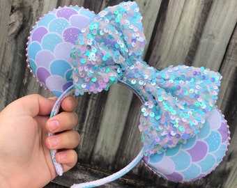 May include: A pair of light blue and purple mouse ears with a large iridescent blue bow. The ears have a mermaid scale pattern.