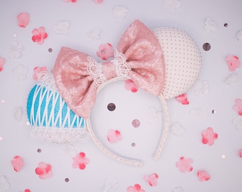 May include: A pair of pink and white mouse ears with a pink bow and a blue and white patterned ear. The ears are on a white background with pink and white confetti.