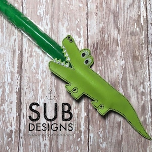 Puede incluir: Una bolsa verde en forma de cocodrilo con una abertura de cremallera, que contiene una paleta de hielo verde en un envoltorio de plástico. La bolsa tiene ojos negros y un diseño cosido.