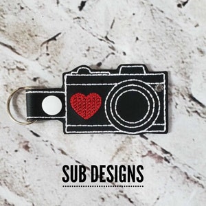 Może przedstawiać: Brelok do kluczy z haftowanym czarno-białym aparatem fotograficznym z czerwonym sercem. Brelok ma biały guzik i srebrny pierścień.