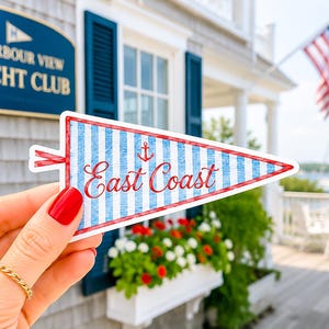 May include: A triangular sticker with a nautical theme, featuring blue and white stripes, a red border, and the words "East Coast" in red script. An anchor graphic is also present. The sticker is held in front of a building with a sign that reads "Harbour View Yacht Club".