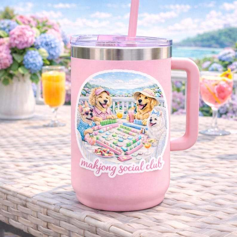 May include: A pink insulated travel mug with a handle and a straw. The mug has a sticker depicting dogs playing mahjong. The text "mahjong social club" is below the image. The background shows a coastal scene.