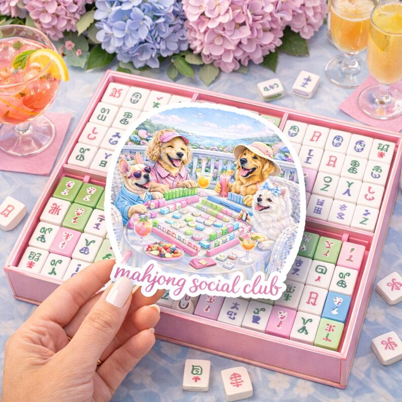 May include: A pink mahjong set with a sticker reading "mahjong social club" and an illustration of dogs playing mahjong. The set is surrounded by drinks and flowers, with tiles in a divided box.