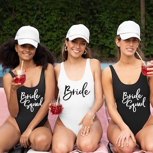 May include: Three women wearing white baseball caps and one-piece swimsuits. Two of the swimsuits are black with white text that says "Bride Squad". The other swimsuit is white with black text that says "Bride".