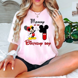 May include: A light pink short-sleeve t-shirt featuring a graphic of Mickey Mouse holding balloons, a large number one shaped like Mickey's head, and the text "Mommy of the Birthday boy" printed in black and red lettering on the front.