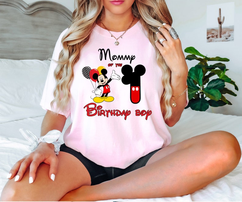 May include: A light pink short-sleeve t-shirt featuring a graphic of Mickey Mouse holding balloons, a large number one shaped like Mickey's head, and the text "Mommy of the Birthday boy" printed in black and red lettering on the front.