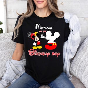 May include: A black short-sleeve t-shirt featuring a graphic of Mickey Mouse holding balloons next to a large number three shaped like Mickey's head. The text reads Mommy of the 3 Birthday boy in white and red lettering on the front.
