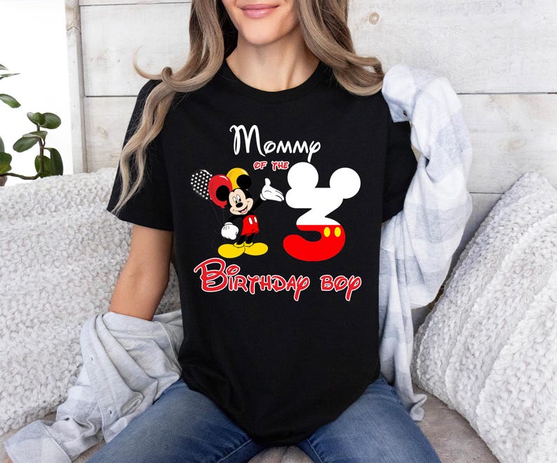 May include: A black short-sleeve t-shirt featuring a graphic of Mickey Mouse holding balloons next to a large number three shaped like Mickey's head. The text reads Mommy of the 3 Birthday boy in white and red lettering on the front.