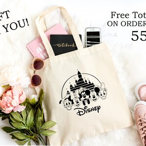 K&ouml;nnte beinhalten: Cremefarbene Tragetasche mit schwarzem Disney-Design, einschlie&szlig;lich Schloss und Figuren. Enth&auml;lt ein rosa Notizbuch, ein schwarzes Notizbuch und ein Smartphone. Der Text lautet "GIFT FOR YOU!" und "Free Tote Bag!" oben.