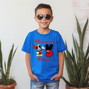 May include: A bright blue short-sleeved t-shirt featuring a graphic of Mickey Mouse holding balloons, the red text "Birthday Boy", a large red number "5" with black mouse ears, and the red name "Bobby" printed on the front of the garment.