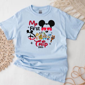 Puede incluir: Camiseta azul claro con un gr&aacute;fico colorido que presenta a Mickey Mouse con gafas de sol, el texto "My First Disney Trip 2026" y otros personajes de Disney. La camiseta es de material suave, ideal para unas vacaciones familiares.