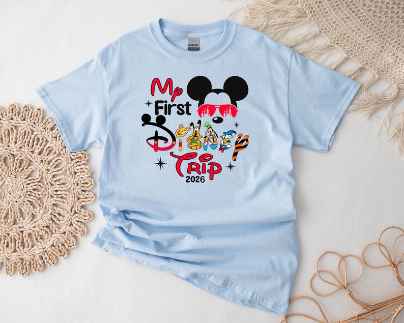 Puede incluir: Camiseta azul claro con un gr&aacute;fico colorido que presenta a Mickey Mouse con gafas de sol, el texto "My First Disney Trip 2026" y otros personajes de Disney. La camiseta es de material suave, ideal para unas vacaciones familiares.