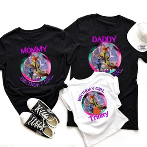 以下が含まれることがあります： 漫画のキャラクターが描かれたカラフルなグラフィックの黒と白のTシャツ。シャツには「Mommy」、「Daddy」、そして「Birthday Girl Trinity」と書かれています。黒いスニーカーと白い帽子も画像にあります。