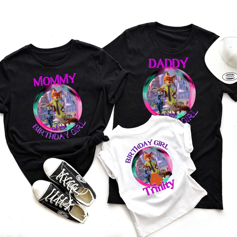 以下が含まれることがあります： 漫画のキャラクターが描かれたカラフルなグラフィックの黒と白のTシャツ。シャツには「Mommy」、「Daddy」、そして「Birthday Girl Trinity」と書かれています。黒いスニーカーと白い帽子も画像にあります。
