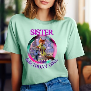 以下が含まれることがあります： Zootopiaのキャラクターのカラフルなグラフィックと「SISTER of the BIRTHAY GIRL」の文字が円でデザインされた、薄い緑色のTシャツ。半袖でクルーネックです。