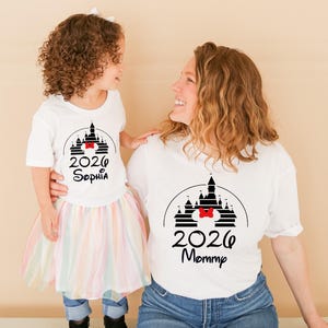K&ouml;nnte beinhalten: Wei&szlig;e T-Shirts mit einem schwarzen Schloss-Design, einer roten Schleife und dem Jahr 2026. Auf einem Shirt steht "Sophia" und auf dem anderen "Mommy". Das Kind tr&auml;gt einen Regenbogen-Tutu, die Erwachsene Jeans.
