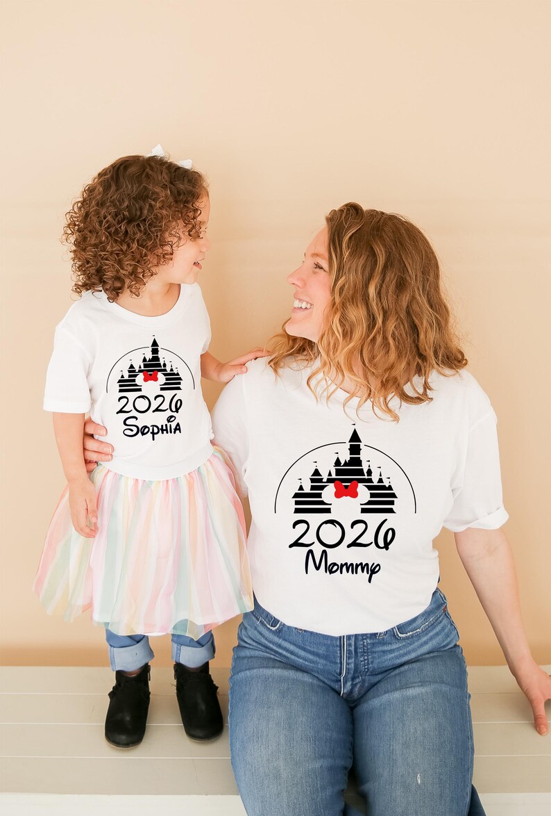 K&ouml;nnte beinhalten: Wei&szlig;e T-Shirts mit einem schwarzen Schloss-Design, einer roten Schleife und dem Jahr 2026. Auf einem Shirt steht "Sophia" und auf dem anderen "Mommy". Das Kind tr&auml;gt einen Regenbogen-Tutu, die Erwachsene Jeans.