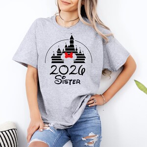 K&ouml;nnte beinhalten: Hellgraues T-Shirt mit schwarzem Schlossdesign, roter Schleife und dem Text "2026 Sister" in Schwarz. Das T-Shirt ist ein l&auml;ssiger Stil, geeignet f&uuml;r den Alltag.