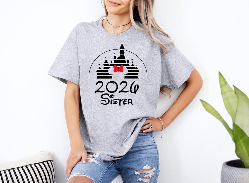 K&ouml;nnte beinhalten: Hellgraues T-Shirt mit schwarzem Schlossdesign, roter Schleife und dem Text "2026 Sister" in Schwarz. Das T-Shirt ist ein l&auml;ssiger Stil, geeignet f&uuml;r den Alltag.