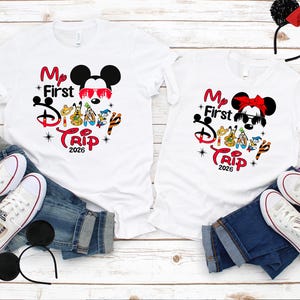 Puede incluir: Dos camisetas blancas con el texto "My First Disney Trip 2026" y gr&aacute;ficos de Mickey y Minnie Mouse. Las camisetas se combinan con vaqueros azules, zapatillas blancas y diademas con orejas de rat&oacute;n. La imagen est&aacute; sobre una superficie de madera clara.
