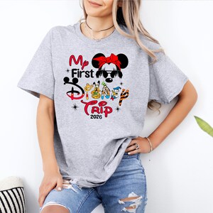 Puede incluir: Camiseta gris claro con un dise&ntilde;o gr&aacute;fico colorido. El dise&ntilde;o incluye el texto "My First Disney Trip 2026" con personajes de Disney y una ilustraci&oacute;n de Minnie Mouse. La camiseta es de manga corta.