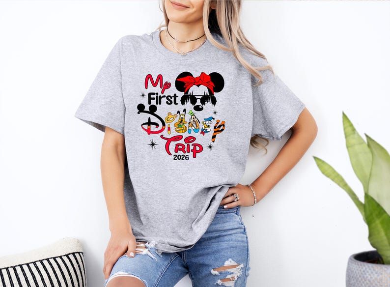 Puede incluir: Camiseta gris claro con un dise&ntilde;o gr&aacute;fico colorido. El dise&ntilde;o incluye el texto "My First Disney Trip 2026" con personajes de Disney y una ilustraci&oacute;n de Minnie Mouse. La camiseta es de manga corta.