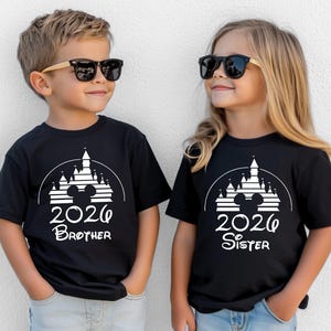 K&ouml;nnte beinhalten: Zwei Kinder tragen schwarze T-Shirts mit einem wei&szlig;en Schloss-Design, Mickey Mouse Ohren und der Jahreszahl 2026. Ein Shirt tr&auml;gt die Aufschrift "Brother", das andere "Sister". Beide Kinder tragen Sonnenbrillen.