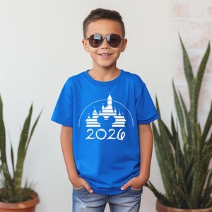 K&ouml;nnte beinhalten: Ein blaues T-Shirt mit einem wei&szlig;en Aufdruck eines Schlosses, Mickey Mouse-Ohren und dem Jahr 2026. Das T-Shirt wird von einer Person mit Sonnenbrille getragen.