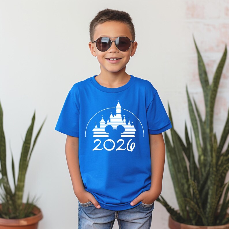 K&ouml;nnte beinhalten: Ein blaues T-Shirt mit einem wei&szlig;en Aufdruck eines Schlosses, Mickey Mouse-Ohren und dem Jahr 2026. Das T-Shirt wird von einer Person mit Sonnenbrille getragen.