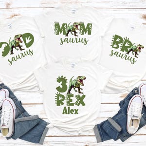 May include: White t-shirts with green dinosaur graphics and text. Designs include "Dad saurus", "Mom saurus", "Bro saurus", and "3 Rex Alex". Paired with blue jeans and white sneakers.