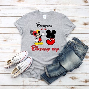 May include: A heather gray short-sleeve t-shirt featuring a graphic of Mickey Mouse holding balloons next to a large number three. The text reads Brother of the Birthday boy. The shirt is paired with blue denim jeans and white canvas sneakers.
