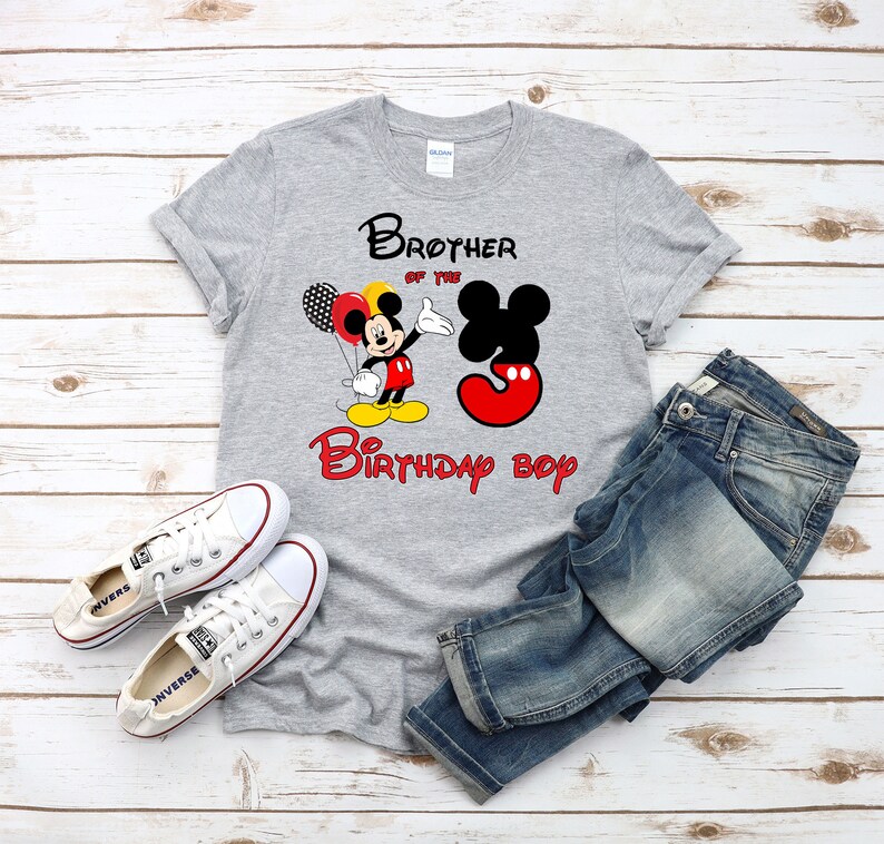May include: A heather gray short-sleeve t-shirt featuring a graphic of Mickey Mouse holding balloons next to a large number three. The text reads Brother of the Birthday boy. The shirt is paired with blue denim jeans and white canvas sneakers.
