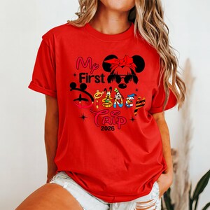 Puede incluir: Camiseta roja con el texto "My First Disney Trip 2026" en rosa, amarillo y negro. El dise&ntilde;o incluye una silueta de Minnie Mouse con un lazo e im&aacute;genes de Goofy, Pato Donald y Pluto.