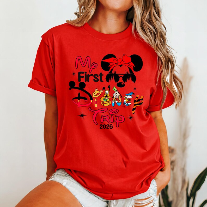 Puede incluir: Camiseta roja con el texto "My First Disney Trip 2026" en rosa, amarillo y negro. El dise&ntilde;o incluye una silueta de Minnie Mouse con un lazo e im&aacute;genes de Goofy, Pato Donald y Pluto.