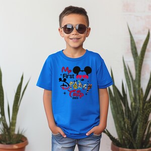 Puede incluir: Una camiseta azul con el texto "My First Disney Trip 2026" en rojo, negro y amarillo. El dise&ntilde;o incluye orejas de Mickey Mouse y otros personajes de Disney. La camiseta la lleva un ni&ntilde;o con gafas de sol.