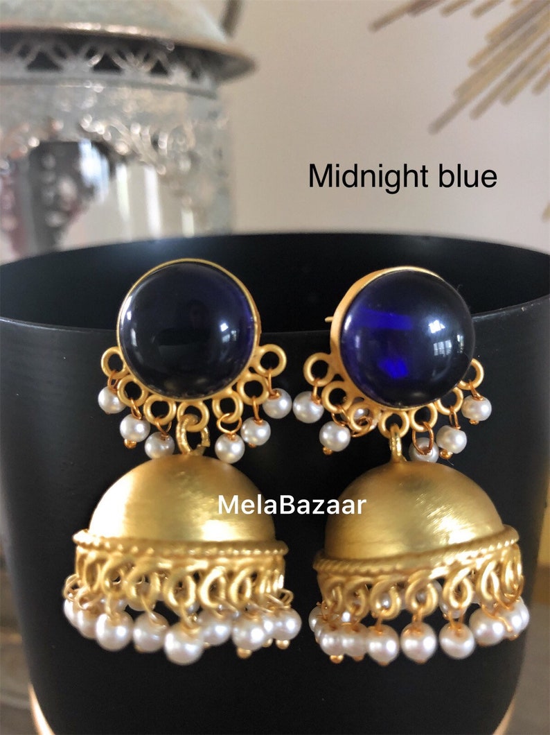 May include: A pair of midnight blue and gold-colored earrings. The earrings have a large, round, dark blue stone at the top, with a gold filigree design. The bottom portion is a gold bell shape with dangling white pearls. The text "Midnight blue" is visible.