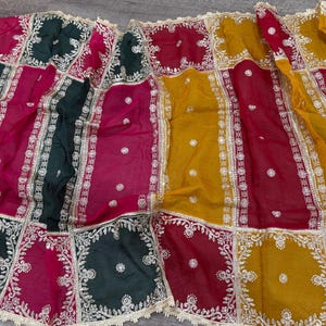 May include: A colorful textile with a patchwork design. The fabric features squares of dark green, magenta, red, and yellow. Each square is bordered with silver embroidery and a delicate gold trim. The textile is likely used for clothing or home decor.