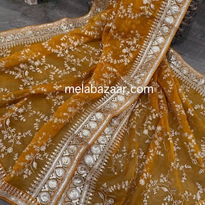 May include: A mustard yellow sheer saree with silver embroidery and sequins. The saree has a floral design and a decorative border.
