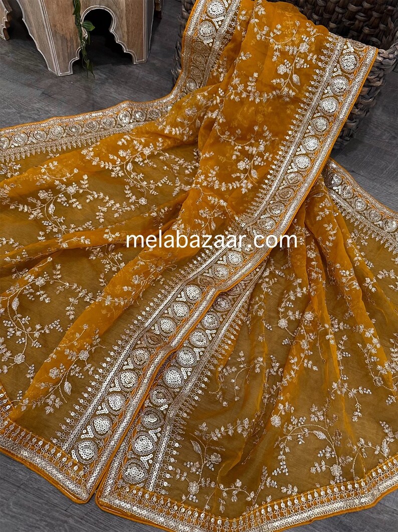 May include: A mustard yellow sheer saree with silver embroidery and sequins. The saree has a floral design and a decorative border.