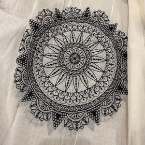 Pode incluir: Close-up de tecido branco com um grande design de mandala bordado em preto. A mandala circular apresenta padr&otilde;es detalhados e um motivo floral central. Bordados florais menores tamb&eacute;m est&atilde;o espalhados pelo tecido.