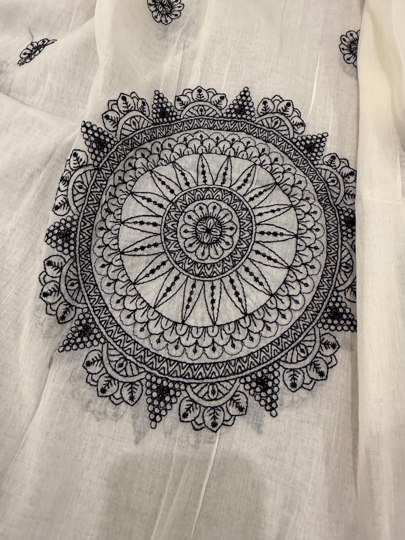 Pode incluir: Close-up de tecido branco com um grande design de mandala bordado em preto. A mandala circular apresenta padr&otilde;es detalhados e um motivo floral central. Bordados florais menores tamb&eacute;m est&atilde;o espalhados pelo tecido.