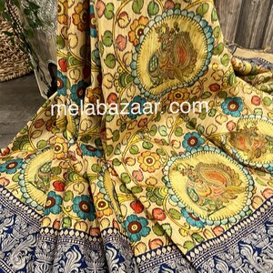 May include: A beige silk saree with a colorful floral and paisley print. The saree has a blue border with a silver design. The saree is folded and displayed on a wooden surface.