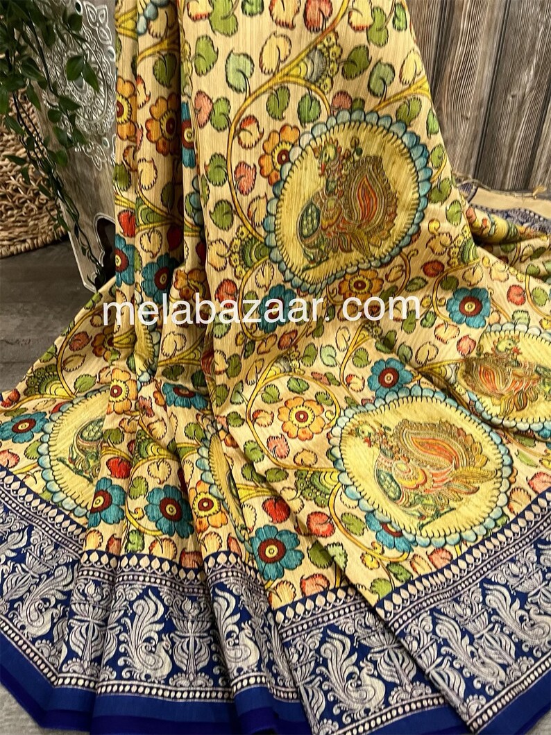 May include: A beige silk saree with a colorful floral and paisley print. The saree has a blue border with a silver design. The saree is folded and displayed on a wooden surface.
