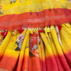 May include: Yellow linen saree with a floral print and a red and orange ombre effect. The saree has a gold trim and tassels.