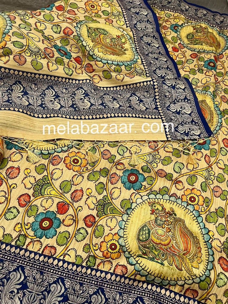 May include: A cream-colored fabric with a blue border and intricate floral and bird designs. The fabric features a repeating pattern of peacocks, flowers, and leaves in shades of blue, green, yellow, red, and orange. The fabric is woven with a fine texture and has a soft, luxurious feel.
