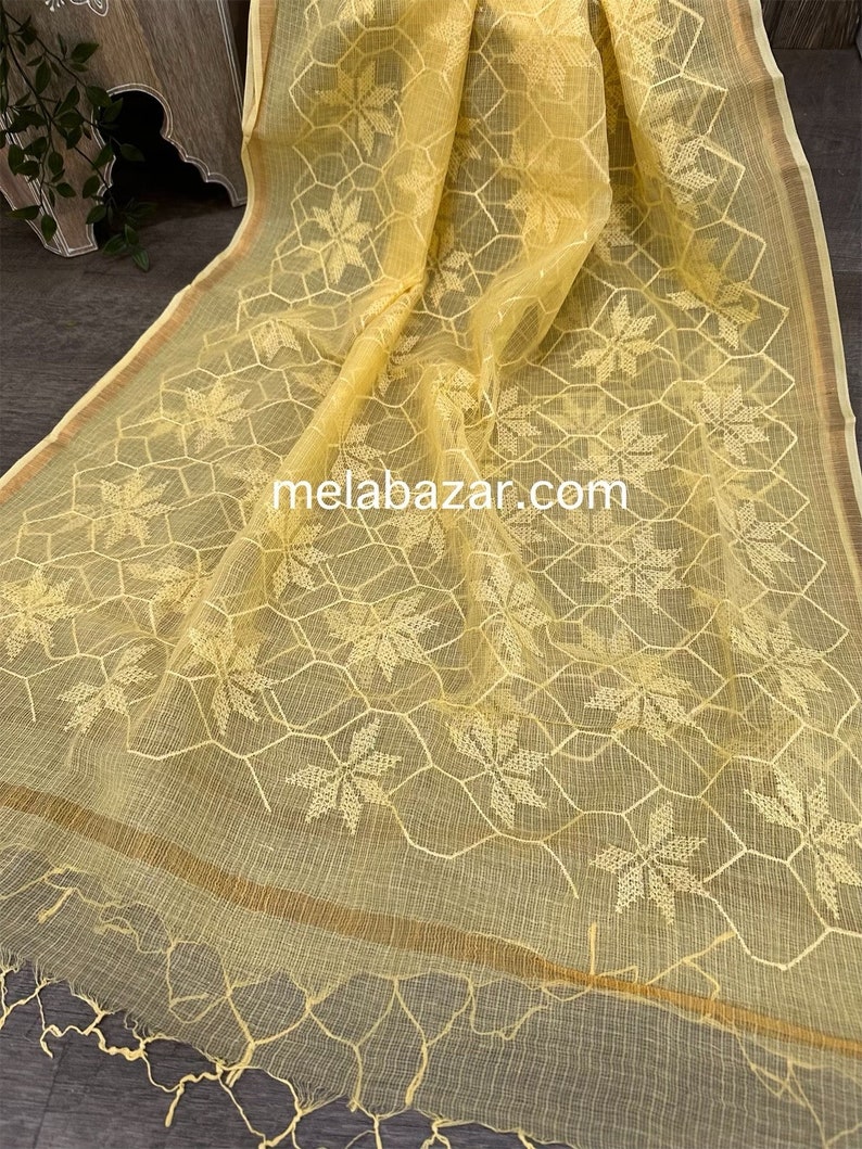 Pode incluir: Um tecido transparente amarelo com um delicado padr&atilde;o bordado de flores e formas geom&eacute;tricas. O tecido tem um brilho sutil e uma textura macia e fluida. O texto "melabazar.com" &eacute; vis&iacute;vel no centro inferior da imagem.
