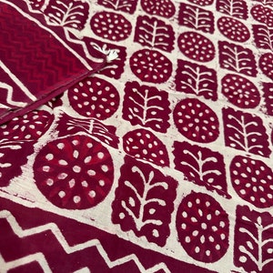 K&ouml;nnte beinhalten: Nahaufnahme eines Textils mit einem sich wiederholenden Muster aus weinroten und wei&szlig;en Formen. Das Design umfasst Kreise, Quadrate und blattartige Formen. Ein weinroter Rand mit Zickzackmuster umrahmt den Stoff. Der Stoff scheint aus Baumwolle zu sein.