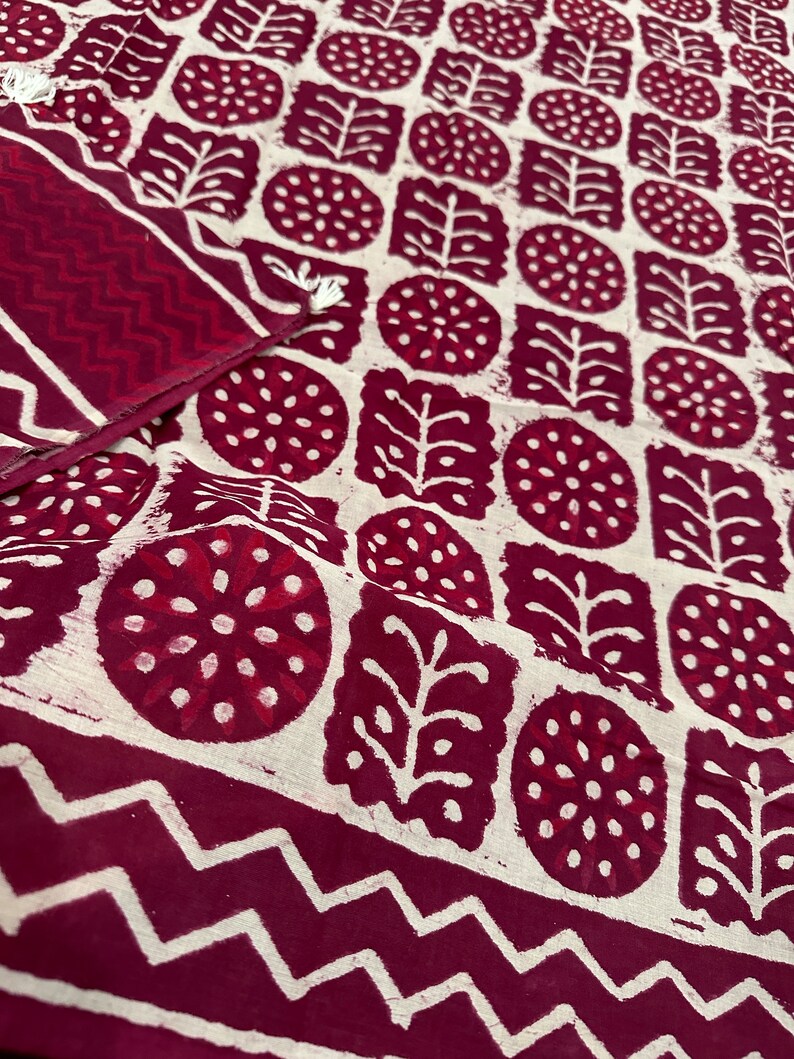 K&ouml;nnte beinhalten: Nahaufnahme eines Textils mit einem sich wiederholenden Muster aus weinroten und wei&szlig;en Formen. Das Design umfasst Kreise, Quadrate und blattartige Formen. Ein weinroter Rand mit Zickzackmuster umrahmt den Stoff. Der Stoff scheint aus Baumwolle zu sein.